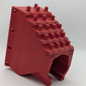 Puede incluir: Una herramienta roja escalonada con clavijas numeradas, diseñada para medir o sujetar objetos. La herramienta tiene los números del 6 al 23 impresos, junto con el texto "3/8"" y "FIW38 M12". La herramienta está hecha de un material sólido y opaco.