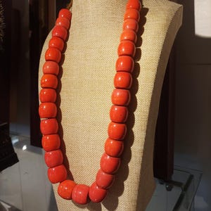 Peut inclure: Un collier de perles rouge vif exposé sur un mannequin. Le collier est composé de grosses perles cylindriques. Les perles sont d'une couleur rouge unie et brillante. Le collier est présenté sur un fond de couleur neutre.