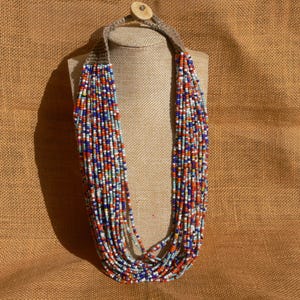 May include: A long multi-strand necklace made of small, colorful beads. The necklace is made of white, blue, red, and orange beads. The necklace is on a brown burlap background.