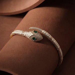 May include: A gold-toned snake bracelet featuring white enamel scales, a head encrusted with small clear stones, and two marquise-cut green stones for the eyes. The flexible metal band wraps around in a coiled design with a textured finish.