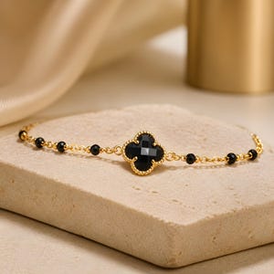 May include: A gold chain bracelet featuring a central four-leaf clover charm with a faceted black stone inlay. The delicate chain is accented with small, round, black beads spaced evenly along the links, creating a refined and balanced design.
