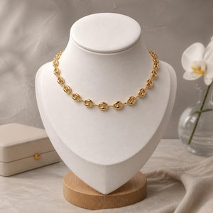 May include: A gold-colored chain necklace with oval links is displayed on a white bust. The necklace is the focal point, showcasing its polished finish. The bust is set on a wooden base, with a small white box and a vase of white flowers in the background.