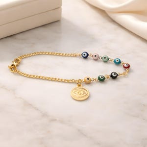 May include: Gold-coloured chain bracelet with colourful beaded eye charms. A gold pendant with an eye design hangs from the chain. The bracelet is displayed on a marble surface.