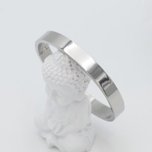 Könnte beinhalten: Ein silberfarbenes, flaches Armreif-Armband. Das Armband wird auf einer weißen Buddha-Statue vor weißem Hintergrund präsentiert. Das Armband hat ein schlichtes, modernes Design.