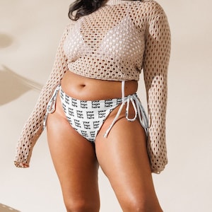 May include: A woman wearing a beige crochet top and a white and black patterned bikini bottom. The bikini bottom has a tie at each side.