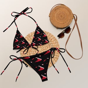 May include: A black and red bikini with a triangle top and a tie-side bottom. The bikini is printed with a pattern of red and white triangles. The bikini is lying on a woven straw mat with a straw bag and sunglasses.