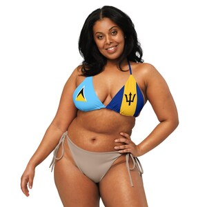 May include: A light blue and yellow bikini top with the flags of Saint Lucia and Barbados. The bikini bottom is a solid taupe color. The woman is smiling and posing with one hand on her hip.