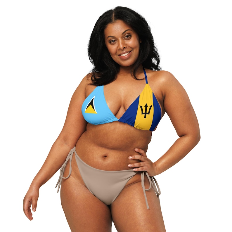 May include: A light blue and yellow bikini top with the flags of Saint Lucia and Barbados. The bikini bottom is a solid taupe color. The woman is smiling and posing with one hand on her hip.