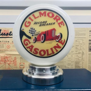 Pode incluir: Um sinal de posto de gasolina de globo branco com um círculo vermelho e amarelo que apresenta um carro de corrida vermelho e o texto "GILMORE the RECORD BREAKER GASOLINE".