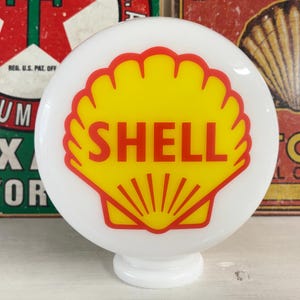Op de afbeelding: Een ronde witte glazen Shell-benzinepompbol met een opvallende gele sint-jakobsschelpgrafiek met een rode omlijning en het woord SHELL in dikke rode hoofdletters gedrukt in het midden van het schelpontwerp op een witte achtergrond.