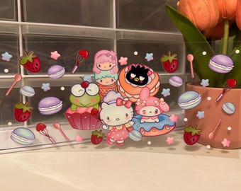 May include: A clear sticker sheet featuring colorful cartoon characters, strawberries, macarons, and cooking utensils. The characters include a frog, a cat, and other cute figures. The overall theme is dessert and kawaii.