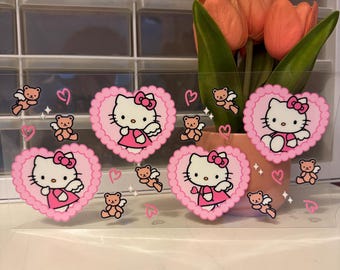 May include: A clear acrylic sheet featuring pink heart-shaped designs with Hello Kitty and teddy bear illustrations. The sheet is decorated with pink hearts and teddy bears with wings. A vase of orange tulips is in the background.