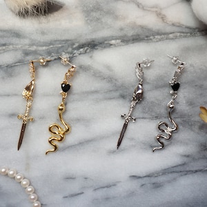 Może przedstawiać: Para kolczyków w kolorze złotym i srebrnym z motywem węża i miecza. Każdy kolczyk ozdobiony jest czarnym serduszkiem.