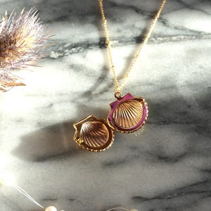 May include: A gold-colored seashell locket necklace with a magenta interior and a gold chain. The locket is open, revealing the interior design. The necklace rests on a marble surface, with a dried flower and a string of pearls in the background.