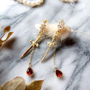 May include: Gold-coloured dagger earrings with a teardrop-shaped, red gemstone pendant. The earrings have a delicate chain and are adorned with small, clear crystals. The background features a marble surface and dried leaves.