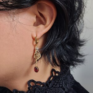 May include: Gold-toned dangle earrings featuring a hand design. The earrings have a small hoop with black stones, a hand pendant with a small diamond-like accent, and a teardrop-shaped red stone at the bottom.