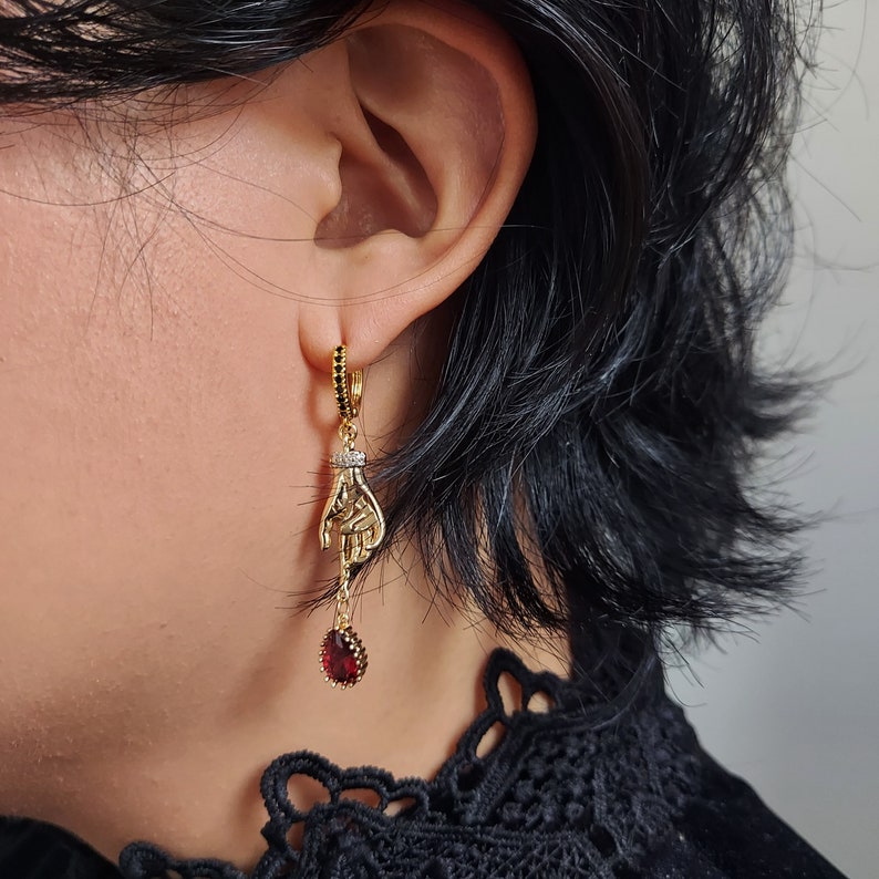 May include: Gold-toned dangle earrings featuring a hand design. The earrings have a small hoop with black stones, a hand pendant with a small diamond-like accent, and a teardrop-shaped red stone at the bottom.