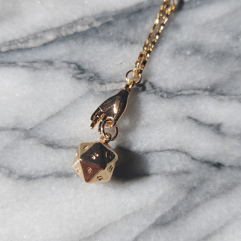May include: Gold necklace with a hand charm holding a gold 20-sided die pendant.
