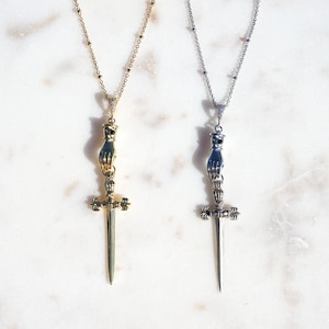 Puede incluir: Dos collares con cadenas delicadas, uno en tono dorado y otro en tono plateado. Cada colgante presenta una mano esculpida que sujeta la empuñadura de una daga puntiaguda. Los detalles de la mano y la empuñadura son intrincados, con una hoja recta.