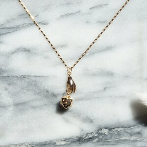 May include: A gold necklace with a hand holding a small gold dice pendant. The necklace is on a white marble background.
