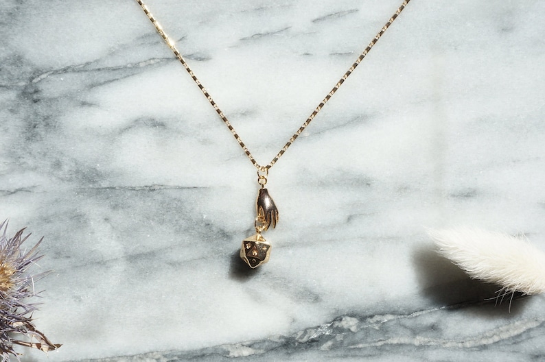May include: A gold necklace with a hand holding a small gold dice pendant. The necklace is on a white marble background.