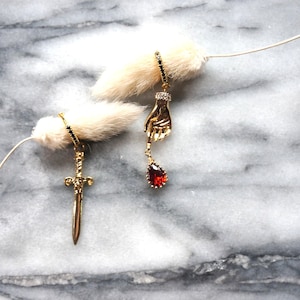 May include: Gold-toned earrings, one with a sword pendant and the other with a hand holding a red teardrop-shaped gem. The earrings are displayed against a marble background with a fluffy white accent.