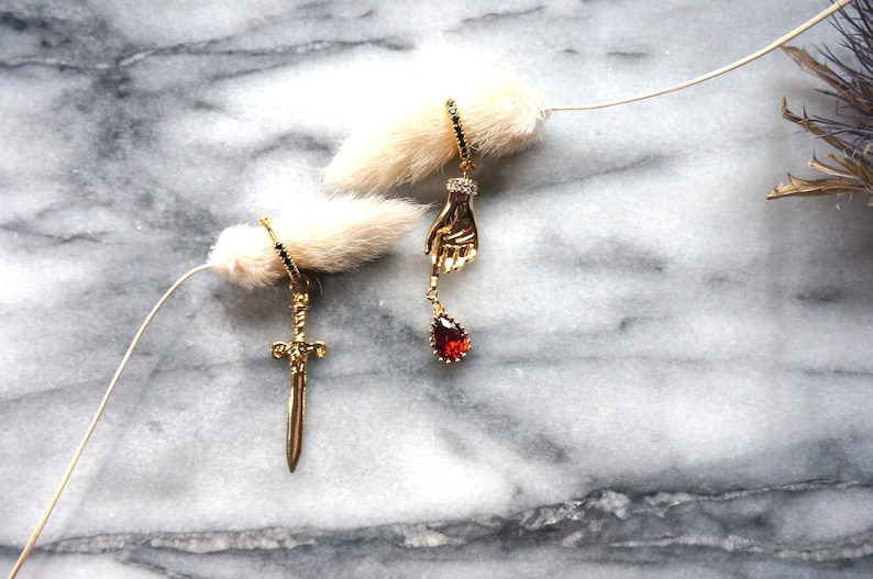 May include: Gold-toned earrings, one with a sword pendant and the other with a hand holding a red teardrop-shaped gem. The earrings are displayed against a marble background with a fluffy white accent.