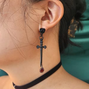 Könnte beinhalten: Schwarze Ohrringe mit schwarzen Rosen-Anhängern und einem schwarzen Dolch mit einem roten Edelstein-Anhänger.