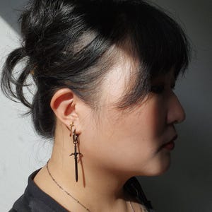 May include: A person with black hair wearing a black shirt and a silver chain necklace with a pendant. They are wearing a black sword-shaped earring in their left ear.