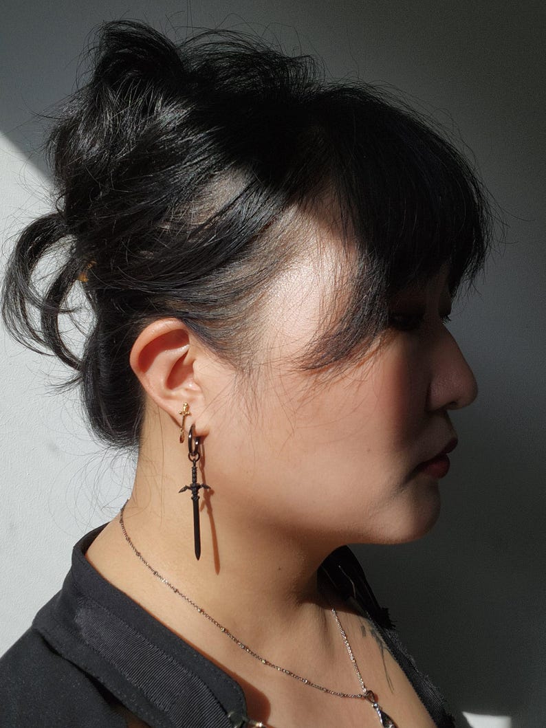 May include: A person with black hair wearing a black shirt and a silver chain necklace with a pendant. They are wearing a black sword-shaped earring in their left ear.