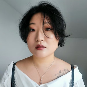 May include: A woman with short black hair wearing a white blouse with a black lace trim and a silver necklace with a small pendant. She is wearing a silver earring on her right ear.