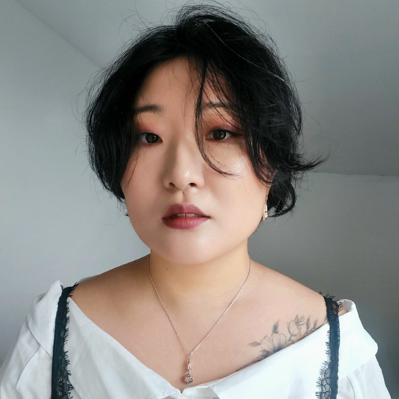 May include: A woman with short black hair wearing a white blouse with a black lace trim and a silver necklace with a small pendant. She is wearing a silver earring on her right ear.