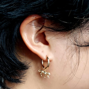 May include: Gold-coloured hoop earring featuring a small leopard charm. The charm has detailed spots and a textured surface, suspended from a simple, polished hoop. The earring is worn on the ear.