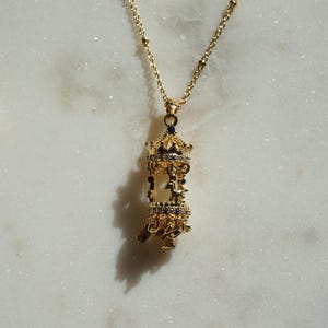 以下が含まれることがあります： 金色のネックレス、メリーゴーランドのペンダント付き。ペンダントは複雑なディテールと小さな輝くアクセントが特徴です。白い大理石の表面に展示されており、白い花の小枝と真珠のネックレスが近くにあります。