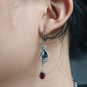 May include: Silver-toned dangle earring featuring a skeletal hand design. The hand holds a teardrop-shaped, deep red gemstone. The earring has a small hoop with black accents and is worn on the ear.