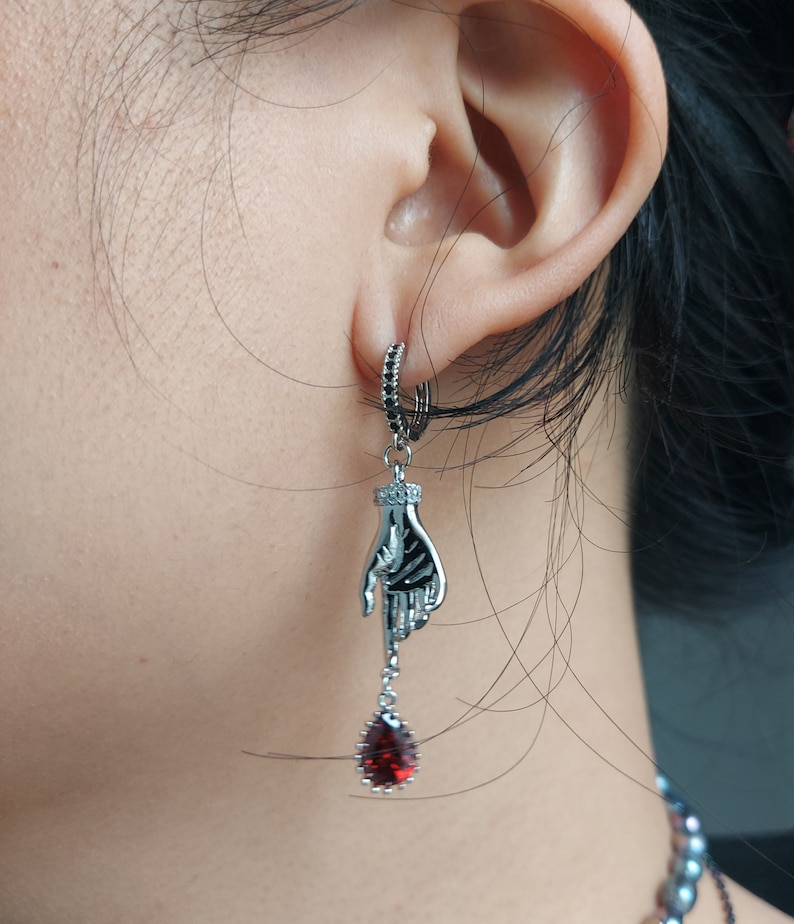 May include: Silver-toned dangle earring featuring a skeletal hand design. The hand holds a teardrop-shaped, deep red gemstone. The earring has a small hoop with black accents and is worn on the ear.