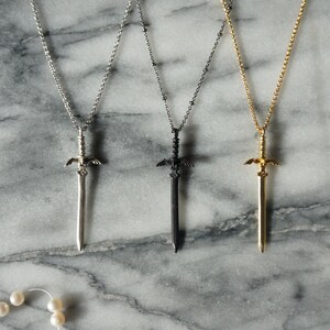 Pu&ograve; includere: Tre collane con ciondoli a forma di spada in argento, nero e oro. Ogni ciondolo presenta un design dettagliato a forma di spada con una catena. Le collane sono esposte su una superficie di marmo, con una collana di perle a sinistra.