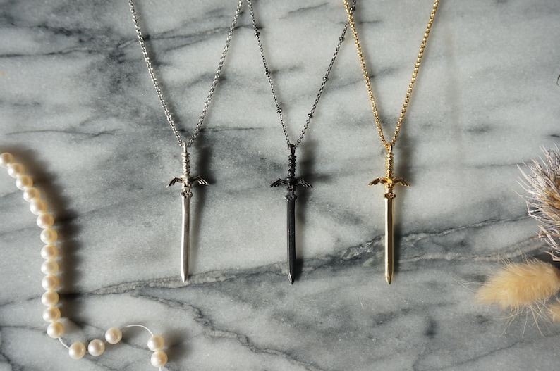 Pu&ograve; includere: Tre collane con ciondoli a forma di spada in argento, nero e oro. Ogni ciondolo presenta un design dettagliato a forma di spada con una catena. Le collane sono esposte su una superficie di marmo, con una collana di perle a sinistra.