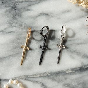 May include: Three sword-shaped earrings with hoop backs. The earrings are gold, black, and silver.