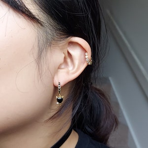 May include: Close-up of an ear featuring two earrings. One is a gold hoop with clear stones. The other is a gold drop earring with a black heart-shaped stone and black stones along the hoop.