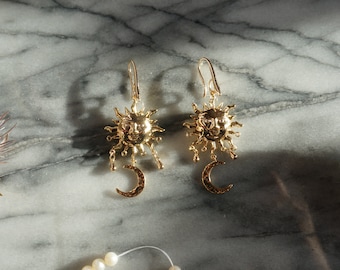 May include: Gold tone earrings featuring a sun and crescent moon design. The sun has a face and rays emanating from it.