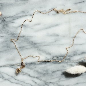 May include: A gold chain necklace with a small, geometric pendant. The pendant features a hand design. The necklace is laying on a marble surface.