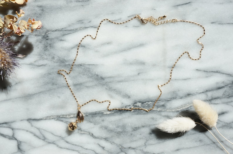 May include: A gold chain necklace with a small, geometric pendant. The pendant features a hand design. The necklace is laying on a marble surface.