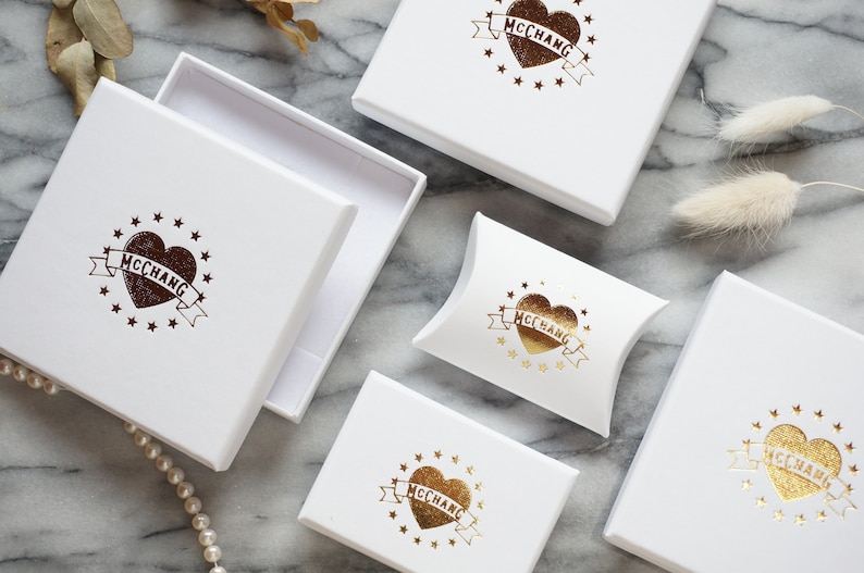 May include: Five white boxes with gold foil logos of a heart with the text "MCCCHANG" inside. The boxes are on a marble surface.