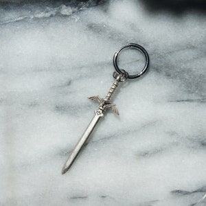 May include: A silver sword charm with a hoop earring attached. The sword has a detailed design with wings and a pointed tip.
