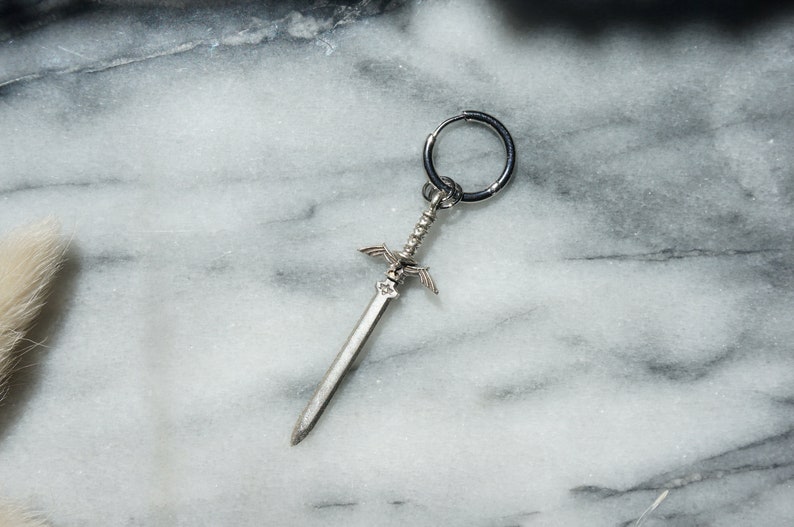 May include: A silver sword charm with a hoop earring attached. The sword has a detailed design with wings and a pointed tip.