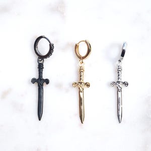 May include: Three hoop earrings with sword-shaped pendants arranged in a row. The first earring is matte black, the second is polished gold-tone, and the third is polished silver-tone. Each sword pendant features a detailed hilt and a straight blade.