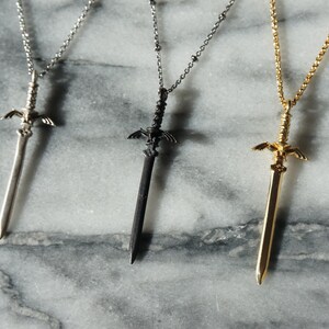 Pu&ograve; includere: Tre collane con ciondoli a forma di spada in argento, nero e oro. Ogni ciondolo presenta un design dettagliato a spada con elsa e ali decorative. Le collane sono esposte su una superficie di marmo.
