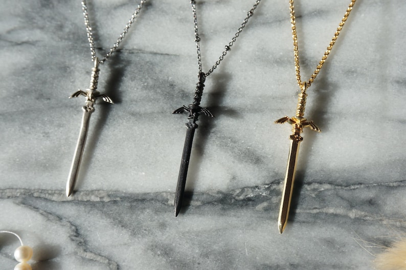 Pu&ograve; includere: Tre collane con ciondoli a forma di spada in argento, nero e oro. Ogni ciondolo presenta un design dettagliato a spada con elsa e ali decorative. Le collane sono esposte su una superficie di marmo.