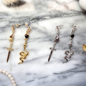 May include: Four pairs of dangle earrings with unique designs. Two pairs are gold-toned, featuring a sword and a snake. The other two pairs are silver-toned, also with a sword and a snake. Each earring has a post back.